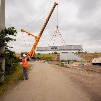 Zdjęcie: na pierwszym planie robotnik, dalej pomarańczowy dźwig podnosi przęsło wiaduktu w Toszku.