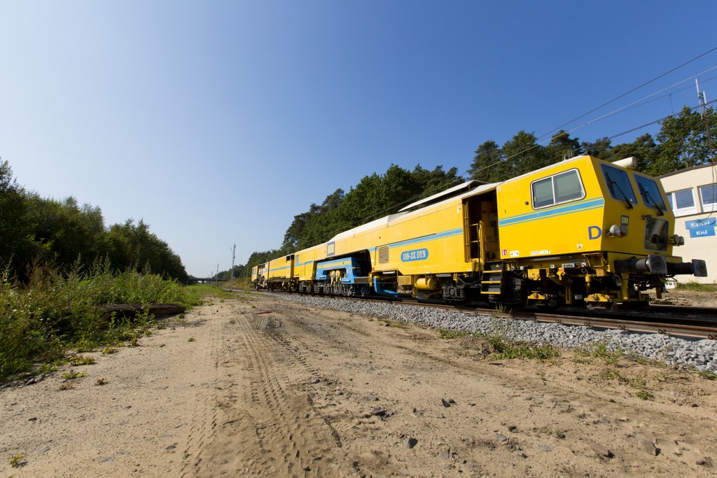 Obrazek: żółty pociąg który jest zgrzewarką torową, w tle las.