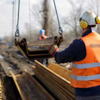 Zdjęcie: robotnik przy pomocy dżwigu umieszcza metalowe wzmocnienie nasypu na ziemi.