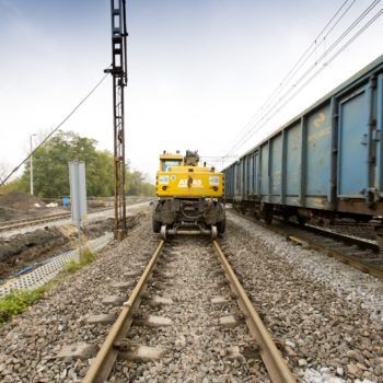 Zdjęcie: koparka porusza się po torach a z prawej strony porusza się pociąg towarowy.