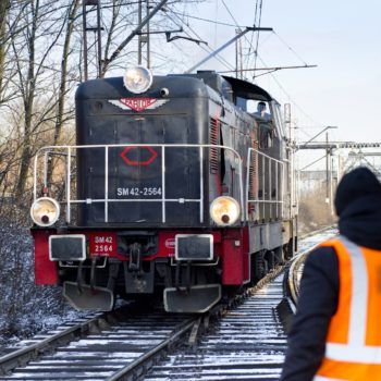 Zdjęcie: starczy czarno czerwony pociąg zbliża się do robotnika pracującego przy podbijarce.