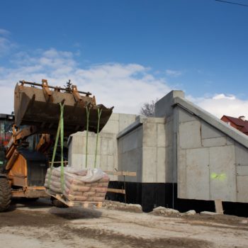 Zdjęcie: koparka wiezie bloki betonowe, w tle betonowa konstrukcja wzmocnienia wiaduktu.