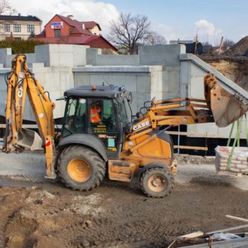 Zdjęcie: koparka wiezie bloki betonowe, w tle betonowa konstrukcja wzmocnienia wiaduktu.