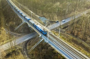 Zdjęcie: widok z góry na wiadukt kolejowo-drogowy przez który jedzie pociąg.