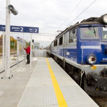 Zdjęcie: podróżni oglądają tablice informacyjną w Chrzanowie. Z lewej jedzie pociąg.