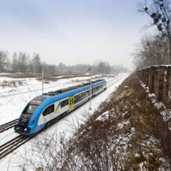 Zdjęcie: w zimowym otoczeniu przejeżdża niebiesko-biały pociąg pasażerski.