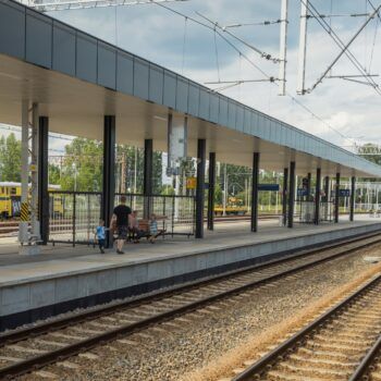 Zdjęcie: widok z poziomu ziemi na świeżo zmodernizowaną stację kolejową Oświęcim. W kadrze widać pasażerów na peronie.
